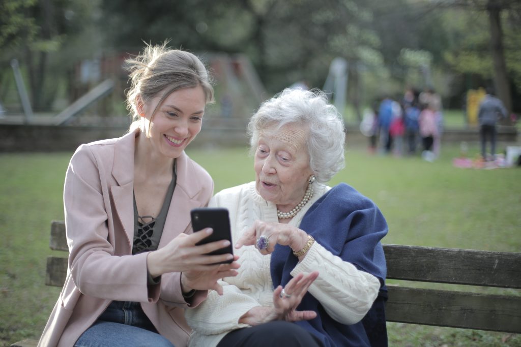 Cheerful senior mother and adult daughter using smartphone together, sitting on a bench at a park. Accessible captions by Subcap.