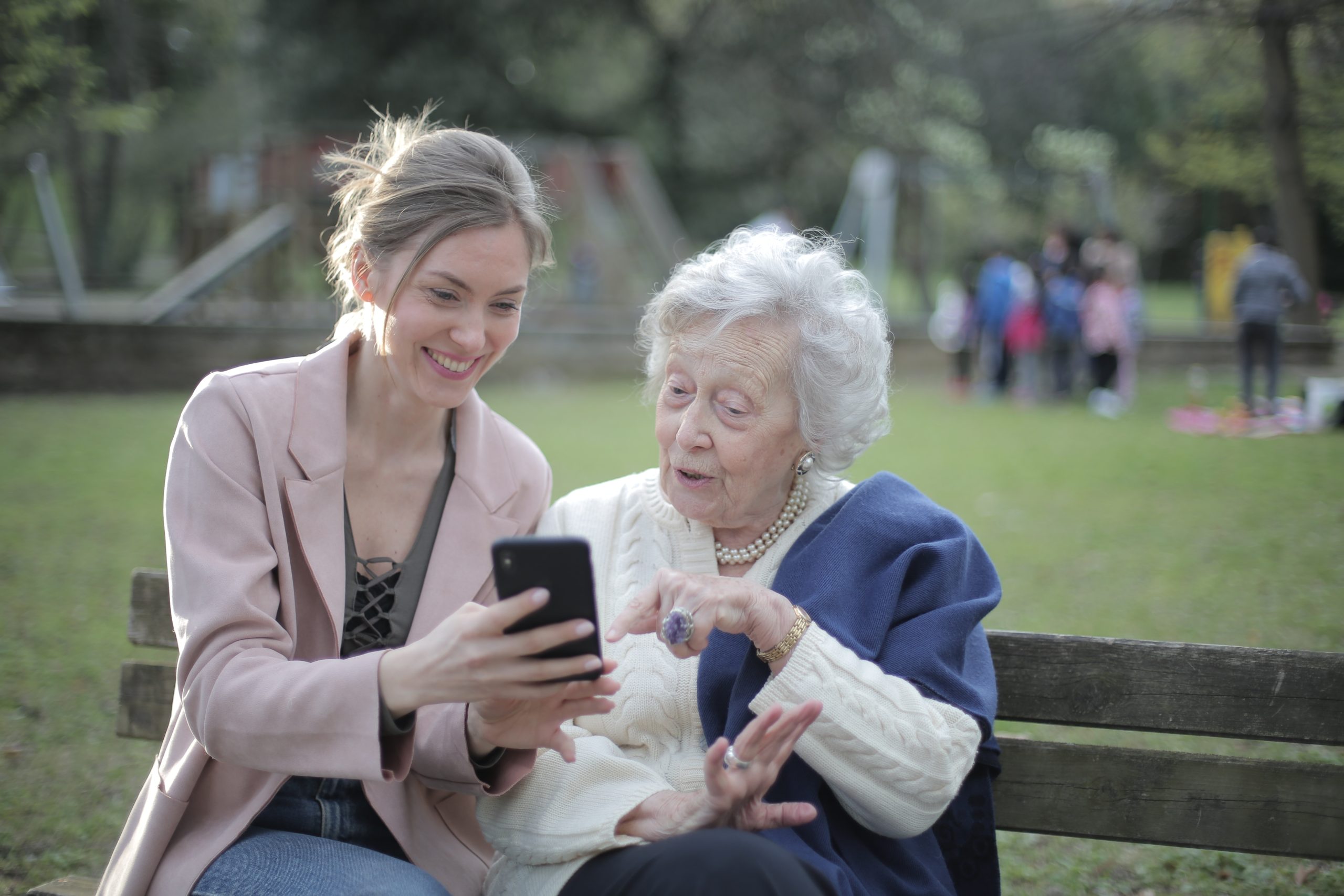 Cheerful senior mother and adult daughter using smartphone together, sitting on a bench at a park